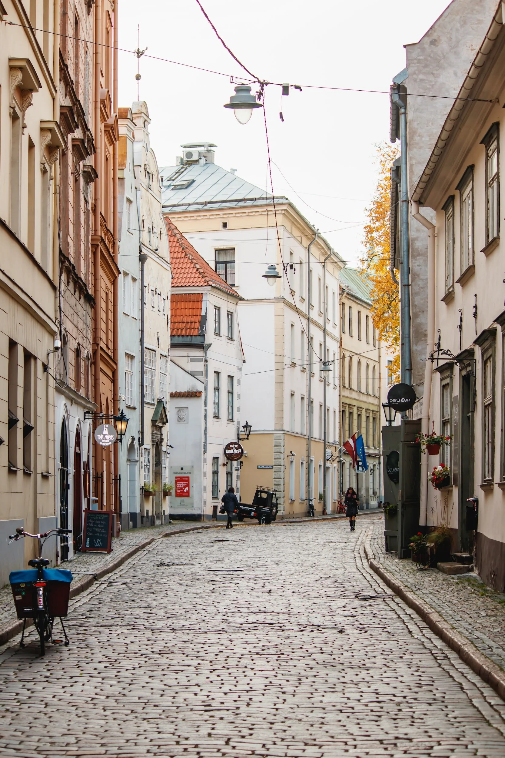 A narrow cobblestone street lined with historic multi-story buildings, some with colorful facades and flower boxes. There are street lamps hanging overhead, a bicycle with a basket on the left sidewalk, and a few pedestrians walking along the street, with some signs and flags visible.