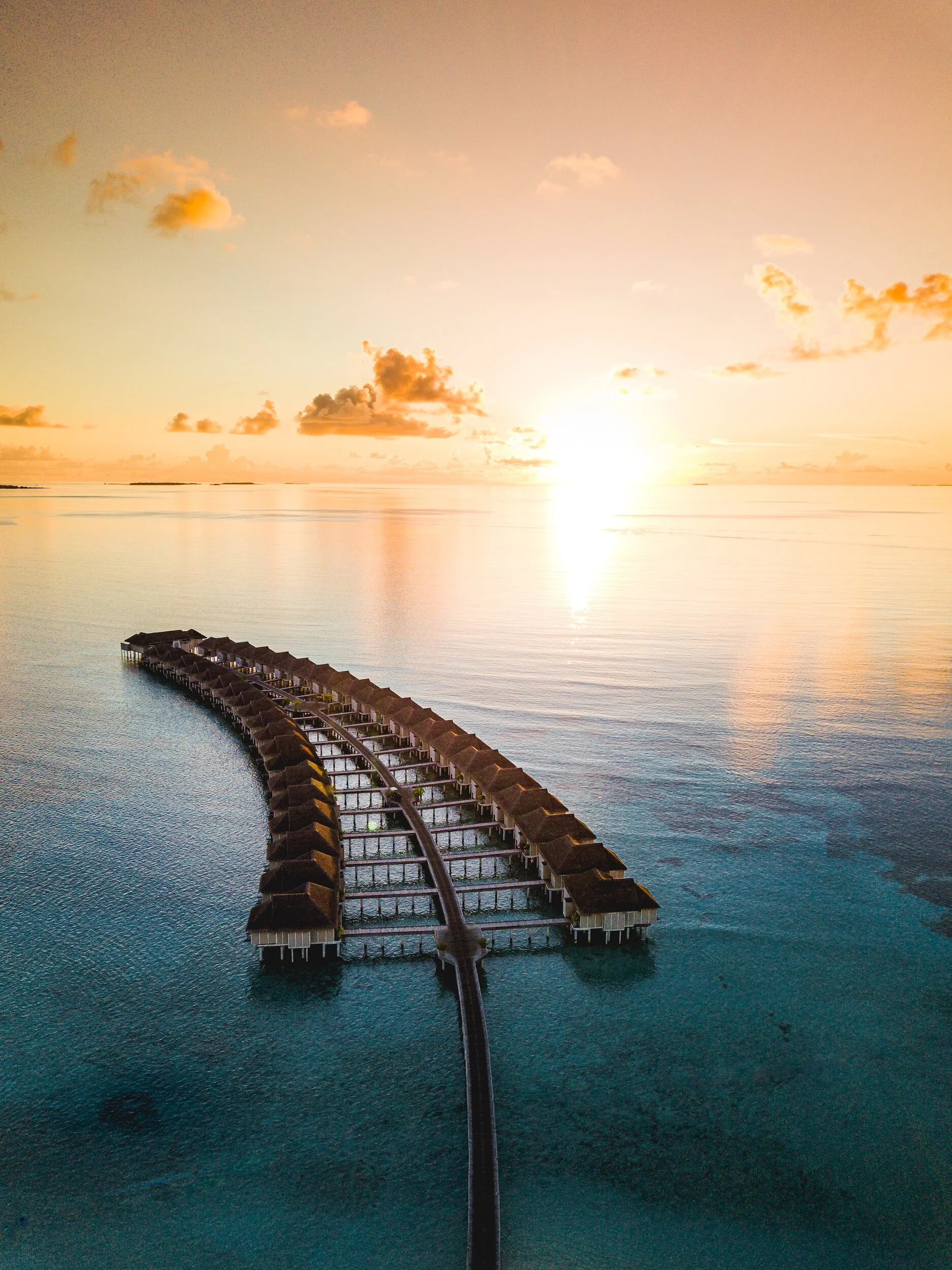 Overwater bungalows on stilts connected by a curved walkway, set in clear ocean water at sunrise or sunset.