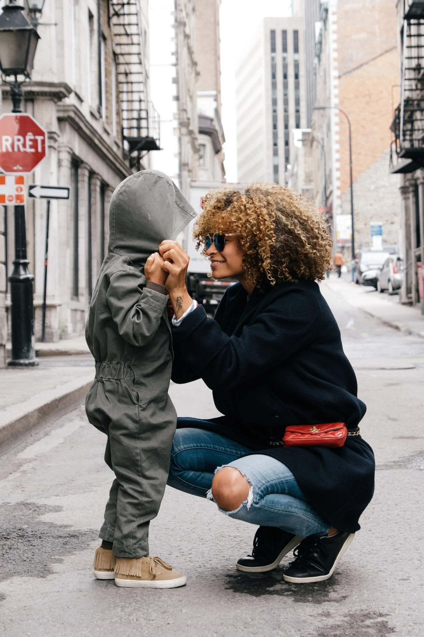 A woman squatting on the street, smiling and holding a child's face close to hers, who is wearing a hooded jacket and tan shoes, in an urban environment with buildings and cars in the background.