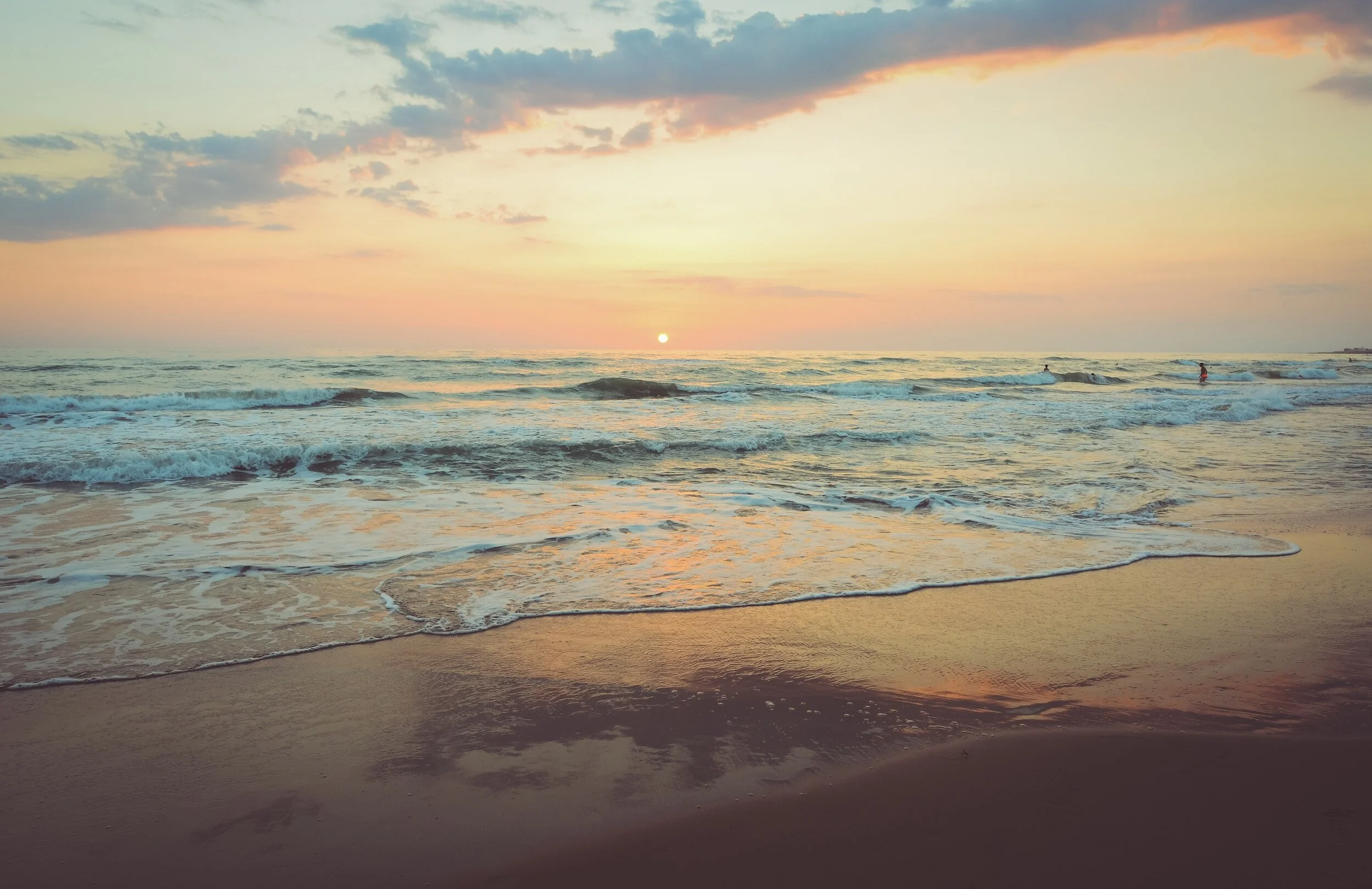 Sunset over the ocean with waves gently crashing on a sandy beach, a few people swimming in the water.